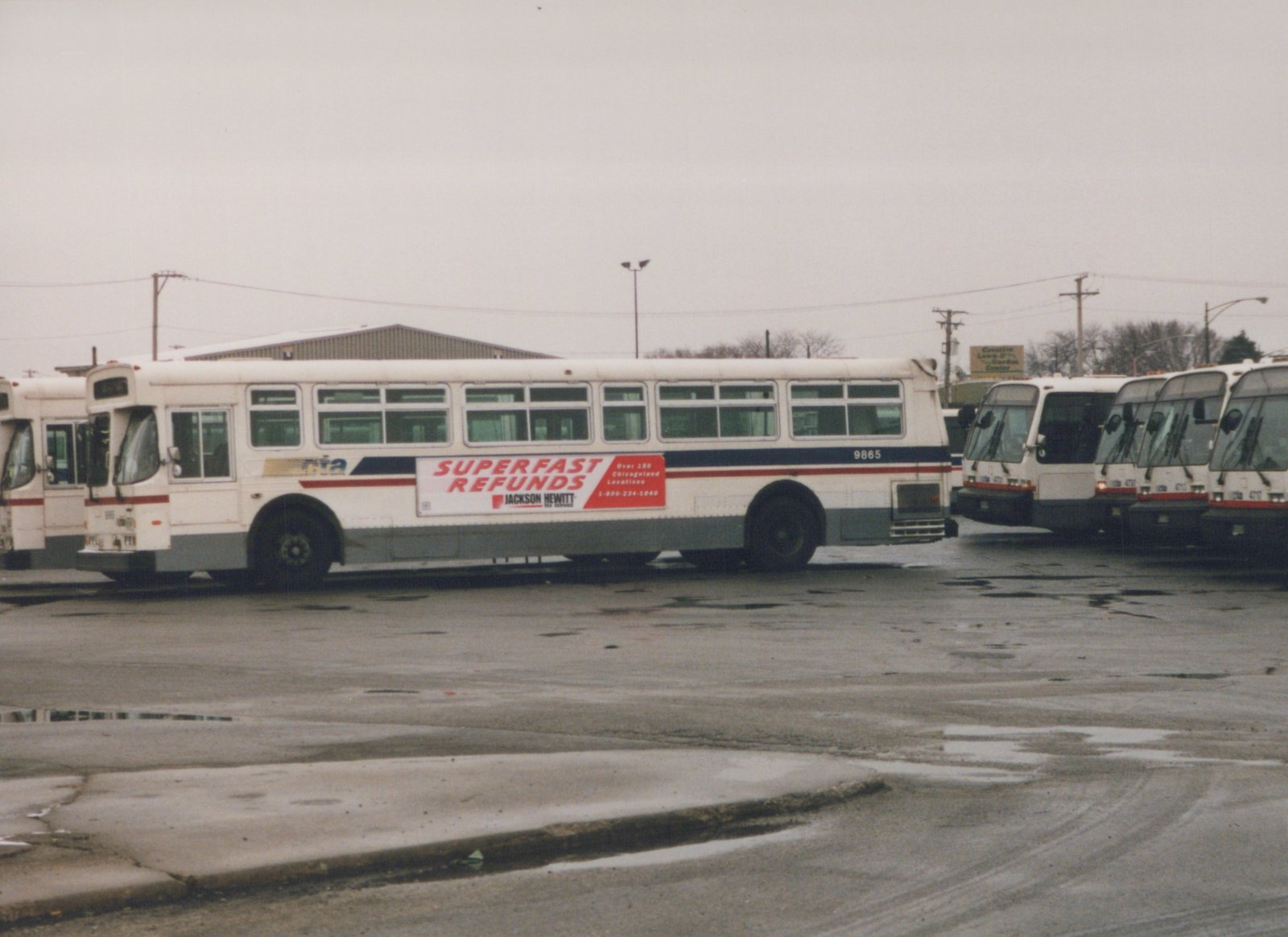 A white CTA bus, number 9865, is parked in a lot with other similar buses under an overcast sky; a Jackson Hewitt ad is on its side.