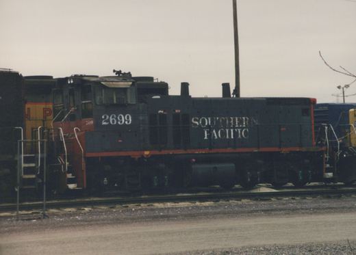 A side view of a dark gray Southern Pacific locomotive, number 2699, sitting on tracks in a yard with other locomotives partially visible behind it.