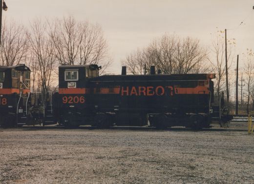Side view of a black and orange Indiana Harbor Belt diesel locomotive numbered 9206, parked on tracks in a gravel yard with bare trees in the background.
