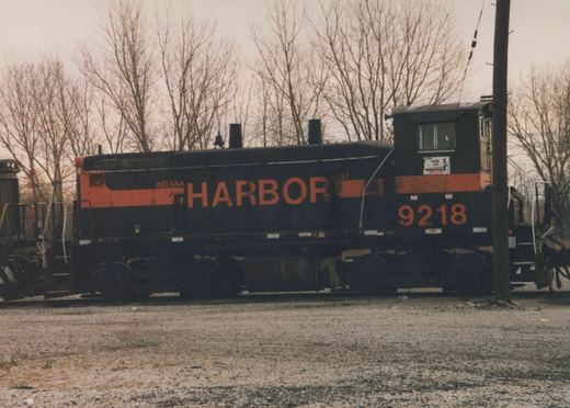 Side view of a black and orange Indiana Harbor Belt diesel locomotive, number 9218, sitting on tracks in a gravel yard with bare trees in the background.