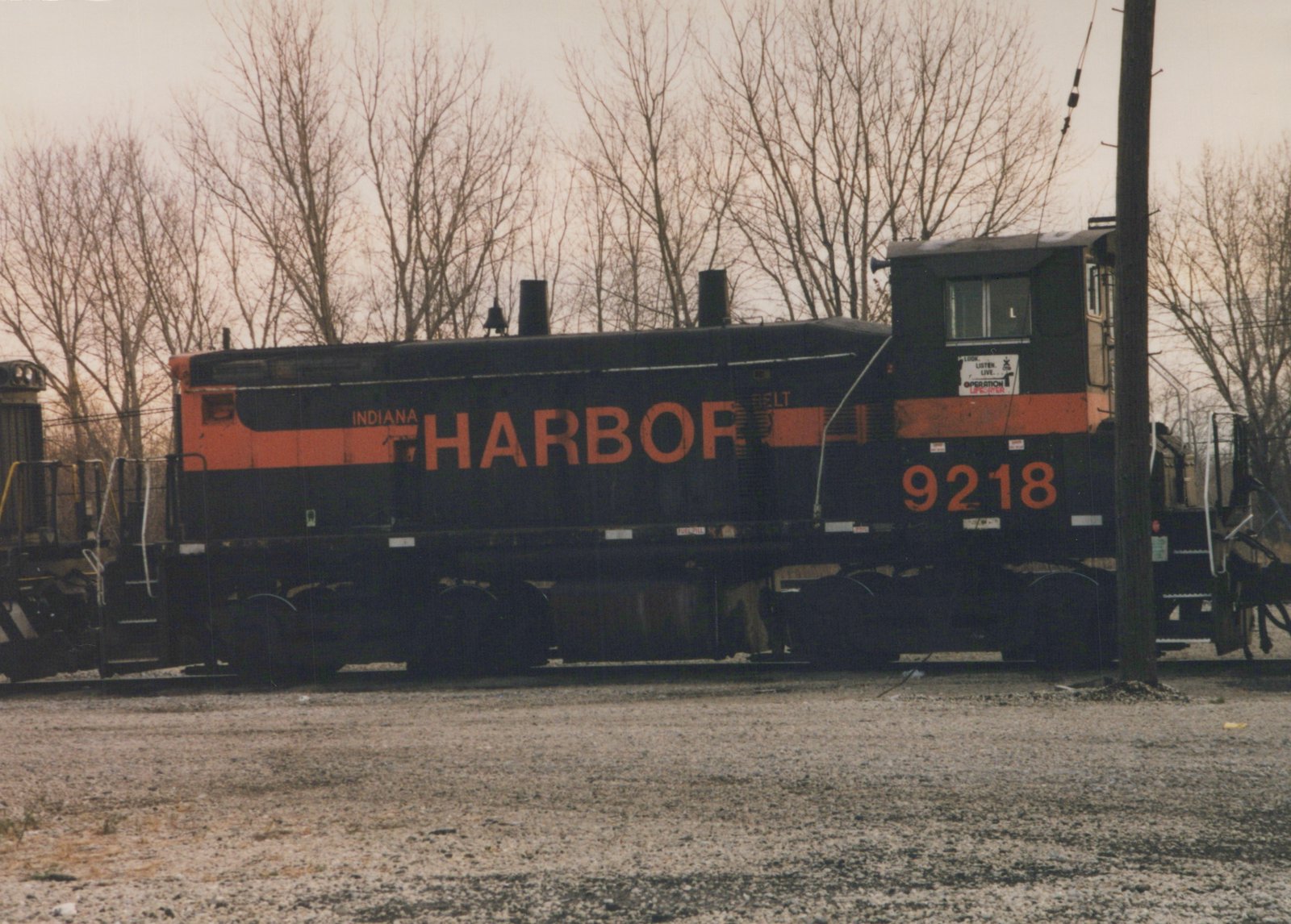 Side view of a black and orange Indiana Harbor Belt diesel locomotive, number 9218, sitting on tracks in a gravel yard with bare trees in the background.