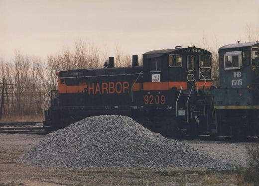 Two diesel locomotives, one black with orange lettering reading 'INDIAN HARBOR 9209' and one dark blue with white lettering reading 'IHB 1505', are coupled in a railroad yard with a pile of gravel and bare trees in the background.