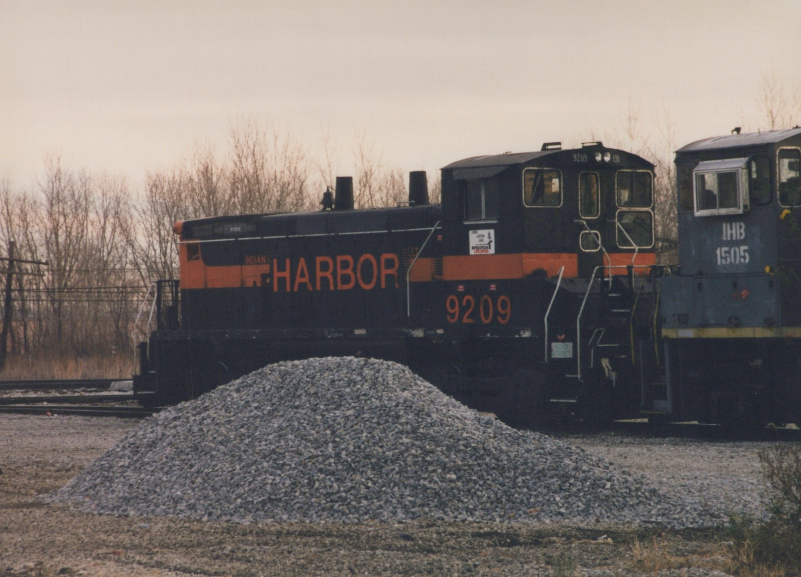 Two diesel locomotives, one black with orange lettering reading 'INDIAN HARBOR 9209' and one dark blue with white lettering reading 'IHB 1505', are coupled in a railroad yard with a pile of gravel and bare trees in the background.