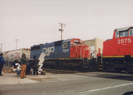 A blue and a red diesel locomotive are coupled on tracks with people nearby holding protest signs, under an overcast sky.