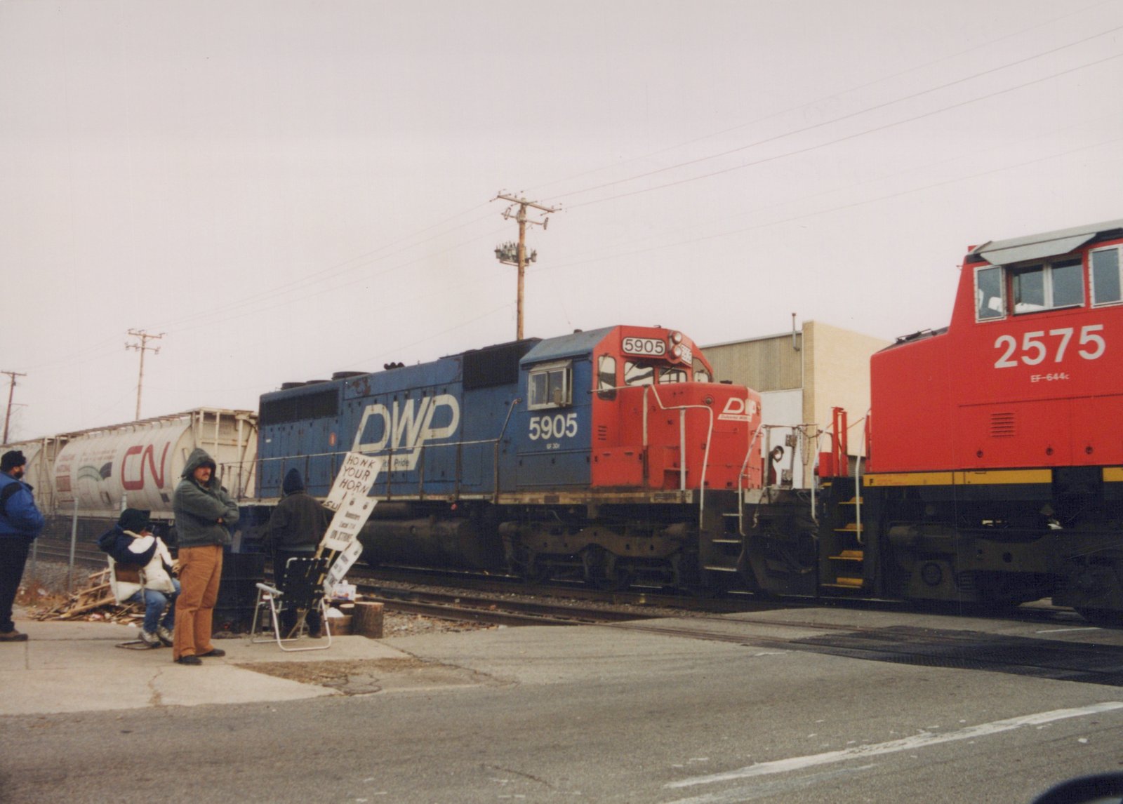 A blue and a red diesel locomotive are coupled on tracks with people nearby holding protest signs, under an overcast sky.