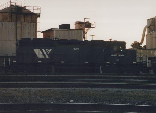 A side view of a black I&M Rail Link diesel locomotive, number 202, sitting on tracks in an industrial yard during dusk.