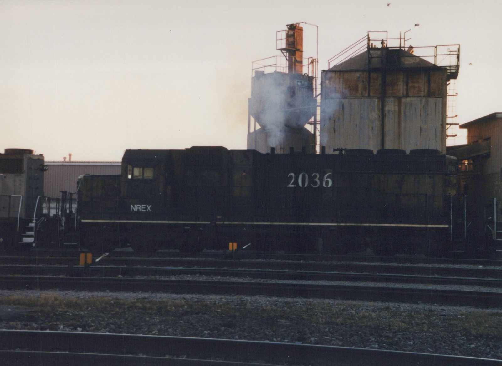 Side view of a dark-colored NREX locomotive numbered 2036 sitting on tracks in a rail yard at dusk, with smoke rising from an industrial building behind it.