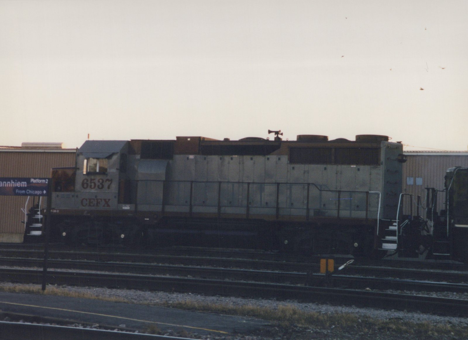 A gray and brown diesel locomotive with the number 6537 and CEFX lettering sits on tracks beside a station platform with a sign reading 'annhiem Platform 2 From Chicago'.
