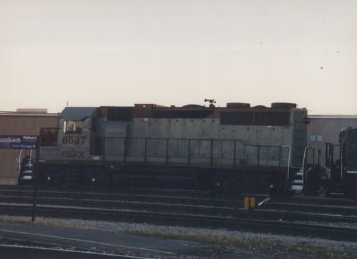 A gray and brown diesel locomotive with the number 6537 and CEFX lettering sits on tracks beside a station platform with a sign reading 'annhiem Platform 2 From Chicago'.