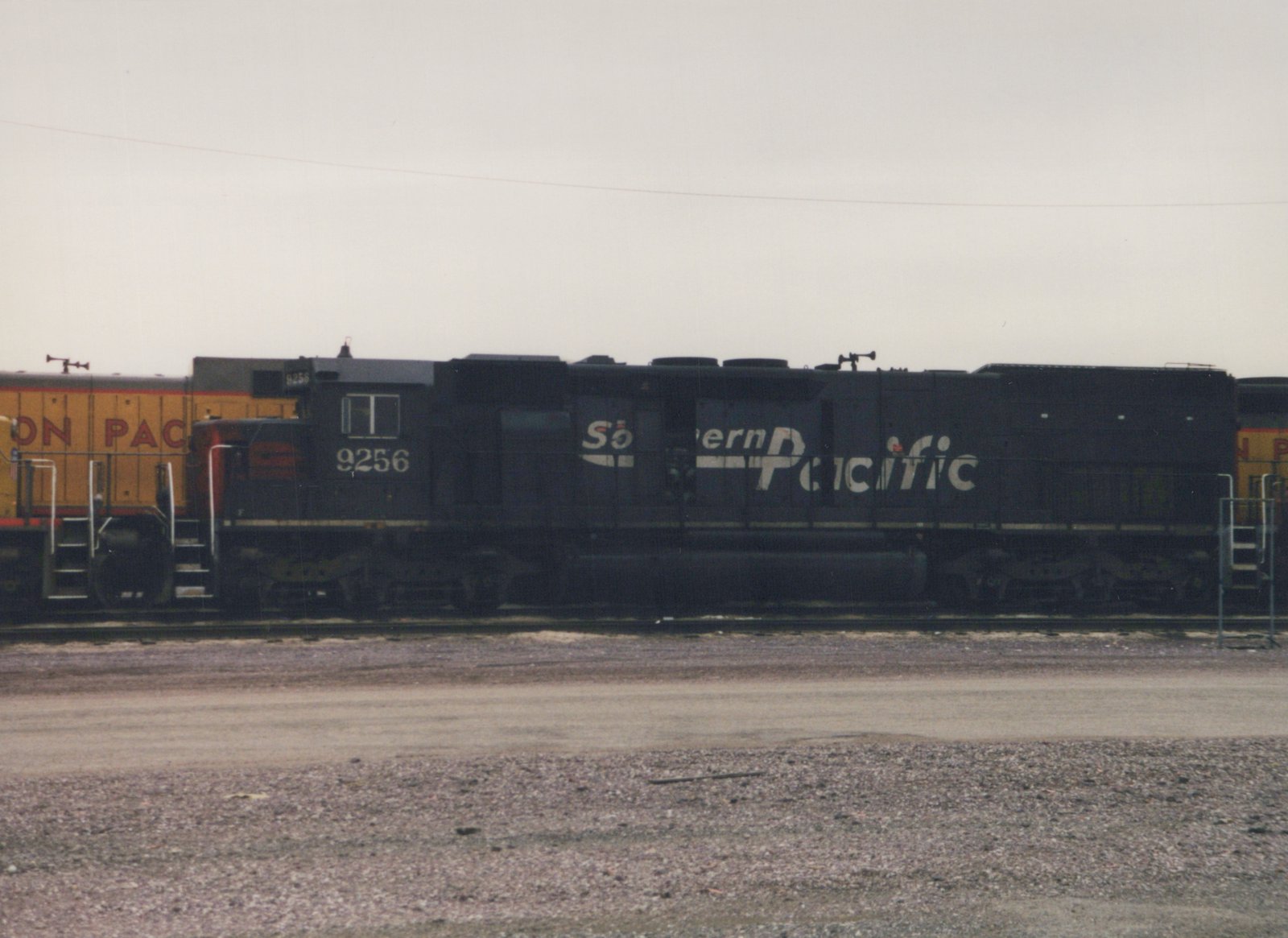 A black Southern Pacific locomotive with the number 9256 is parked on a set of tracks in a gravel yard, with a yellow locomotive visible in the background.