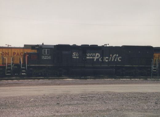 A black Southern Pacific locomotive with the number 9256 is parked on a set of tracks in a gravel yard, with a yellow locomotive visible in the background.