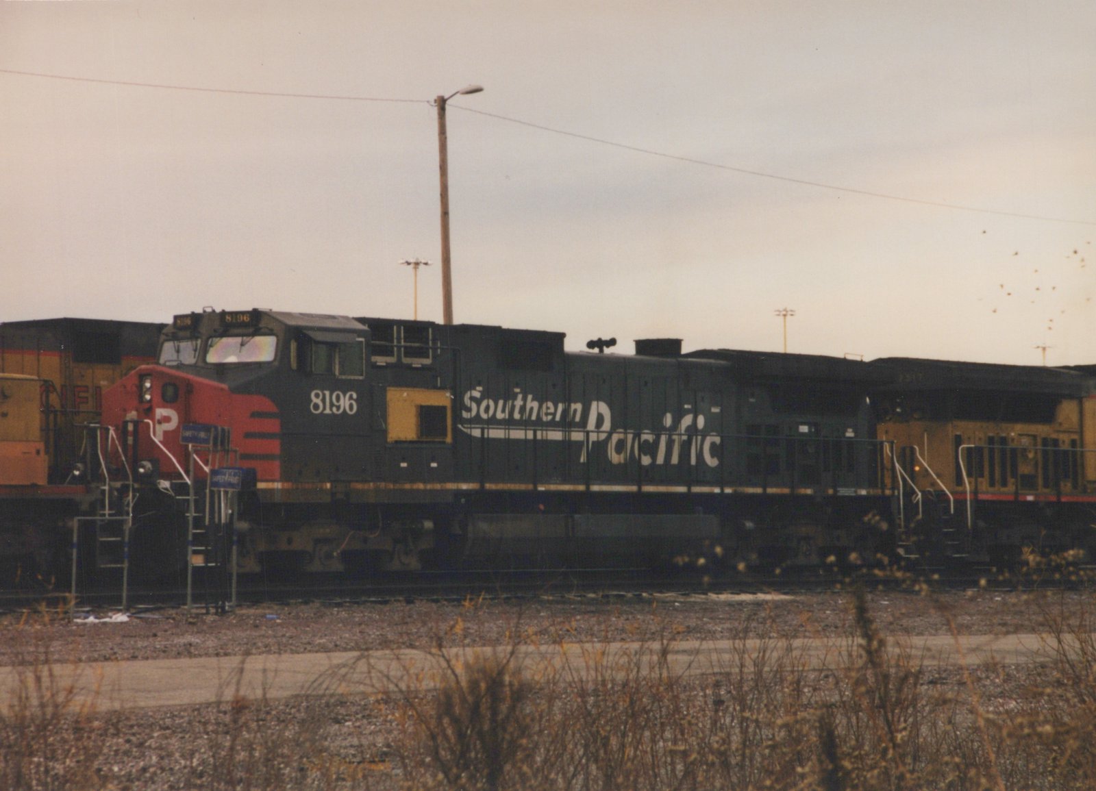 A black Southern Pacific diesel locomotive numbered 8196 is shown from the side, stopped in a rail yard under an overcast sky. Another locomotive is visible behind it.