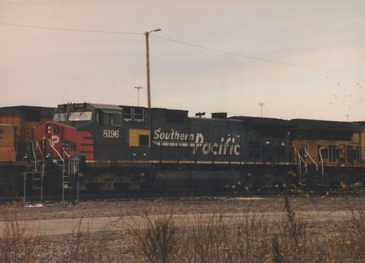 A black Southern Pacific diesel locomotive numbered 8196 is shown from the side, stopped in a rail yard under an overcast sky. Another locomotive is visible behind it.