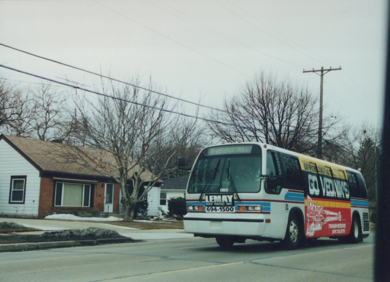 A white Lemay Flxible Metro bus, number 065, drives down a snow-dusted residential street under a gray sky, with bare trees and houses in the background.