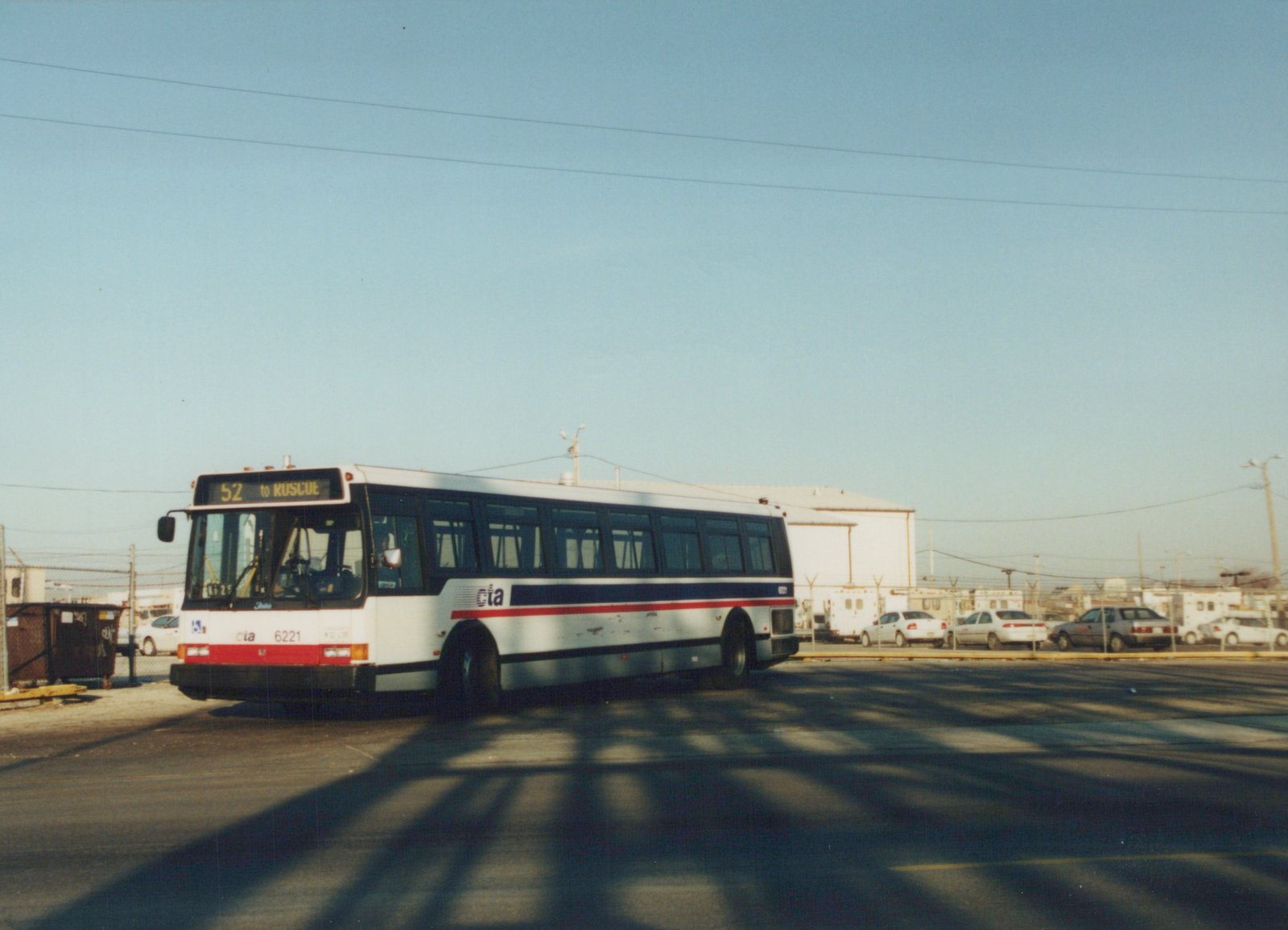 A white Chicago Transit Authority bus with blue and red stripes, number 6221, is parked on a street with a clear sky and several cars in the background.