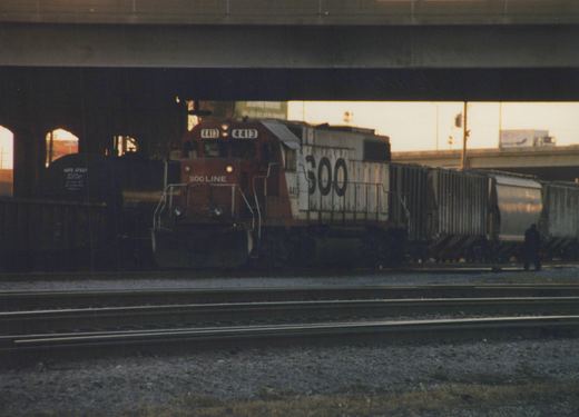 A maroon and white Soo Line diesel locomotive numbered 4413 is shown pulling a long line of freight cars on railroad tracks beneath a large concrete overpass during the evening.