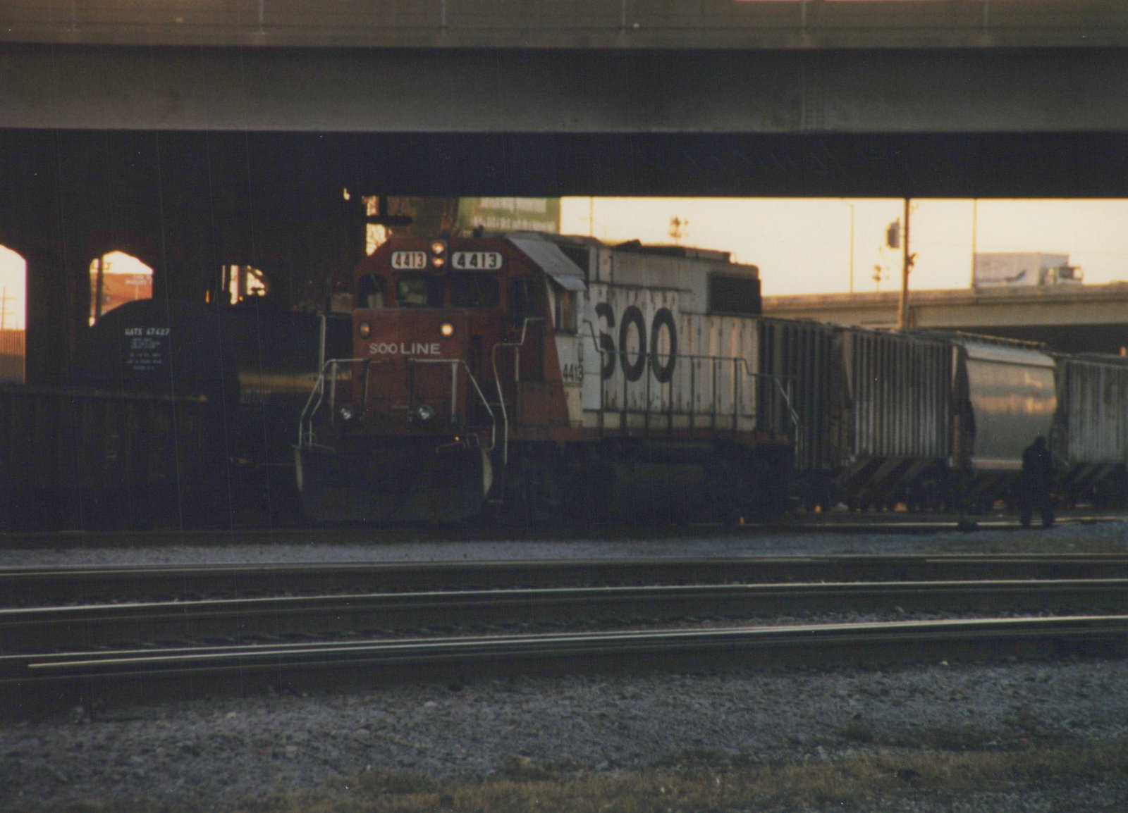 A maroon and white Soo Line diesel locomotive numbered 4413 is shown pulling a long line of freight cars on railroad tracks beneath a large concrete overpass during the evening.