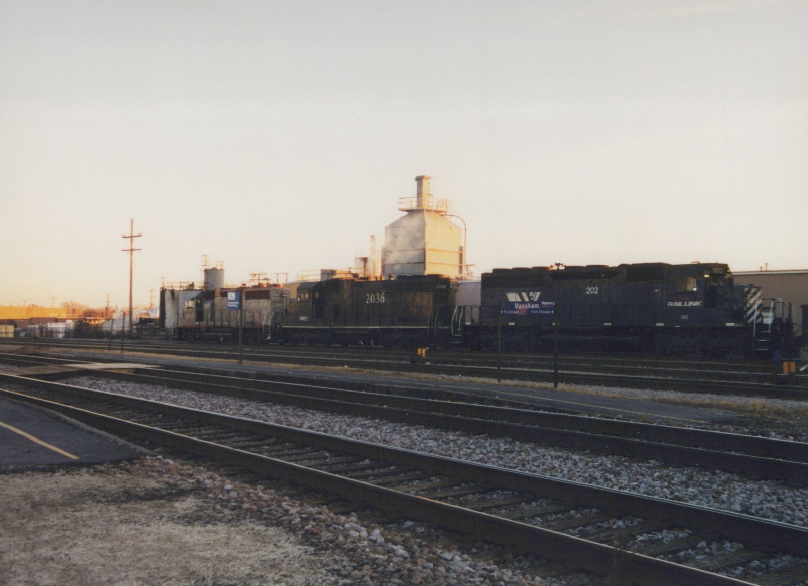 Two diesel locomotives, one blue with 'Rail Link' lettering and number 202, and one dark with number 2036, sit on parallel tracks at a railroad yard at dusk.