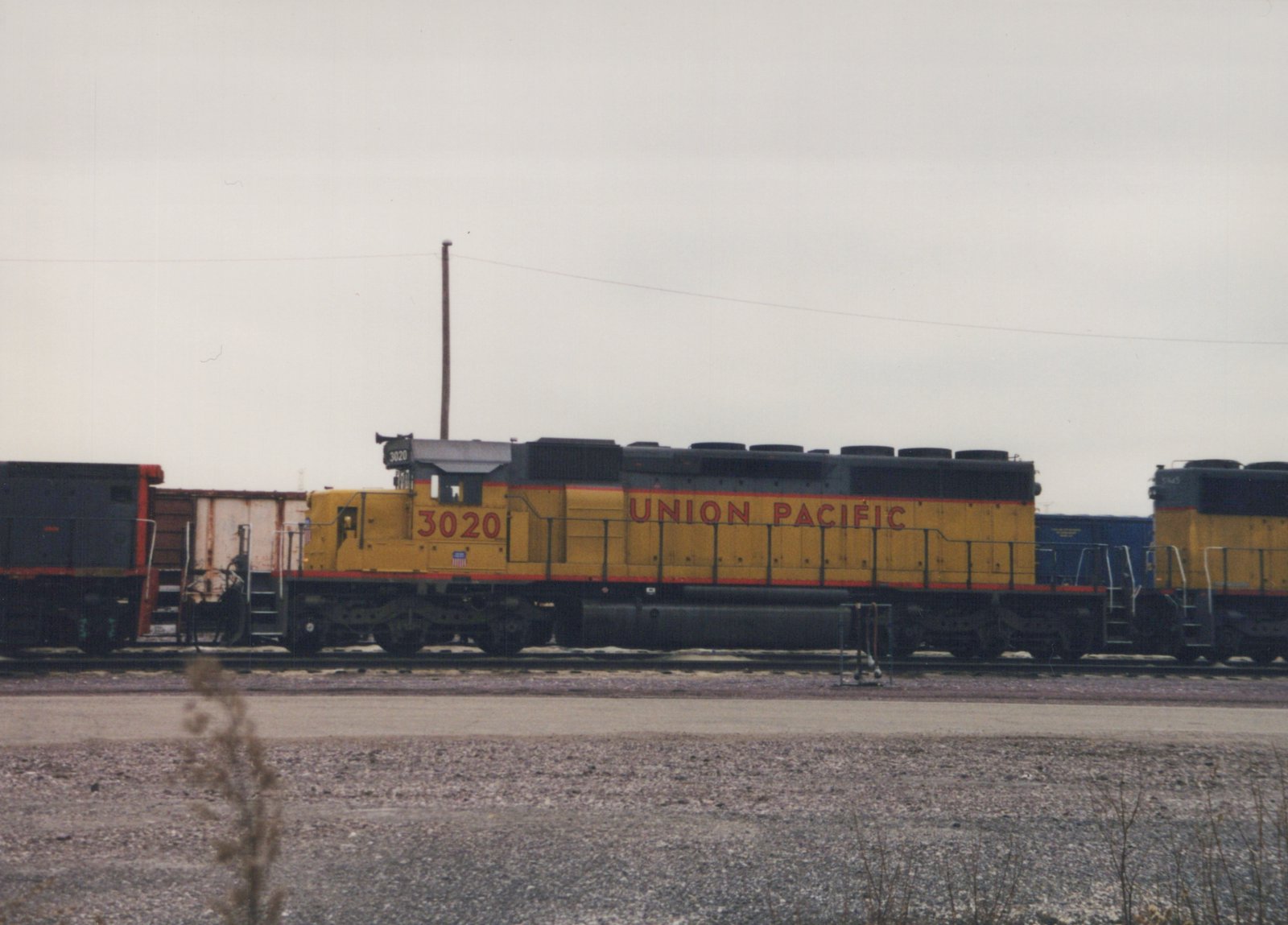 Side view of a yellow and black Union Pacific diesel locomotive with the number 3020, standing in a gravel rail yard under a grey sky.