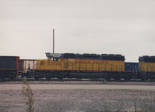 Side view of a yellow and black Union Pacific diesel locomotive with the number 3020, standing in a gravel rail yard under a grey sky.