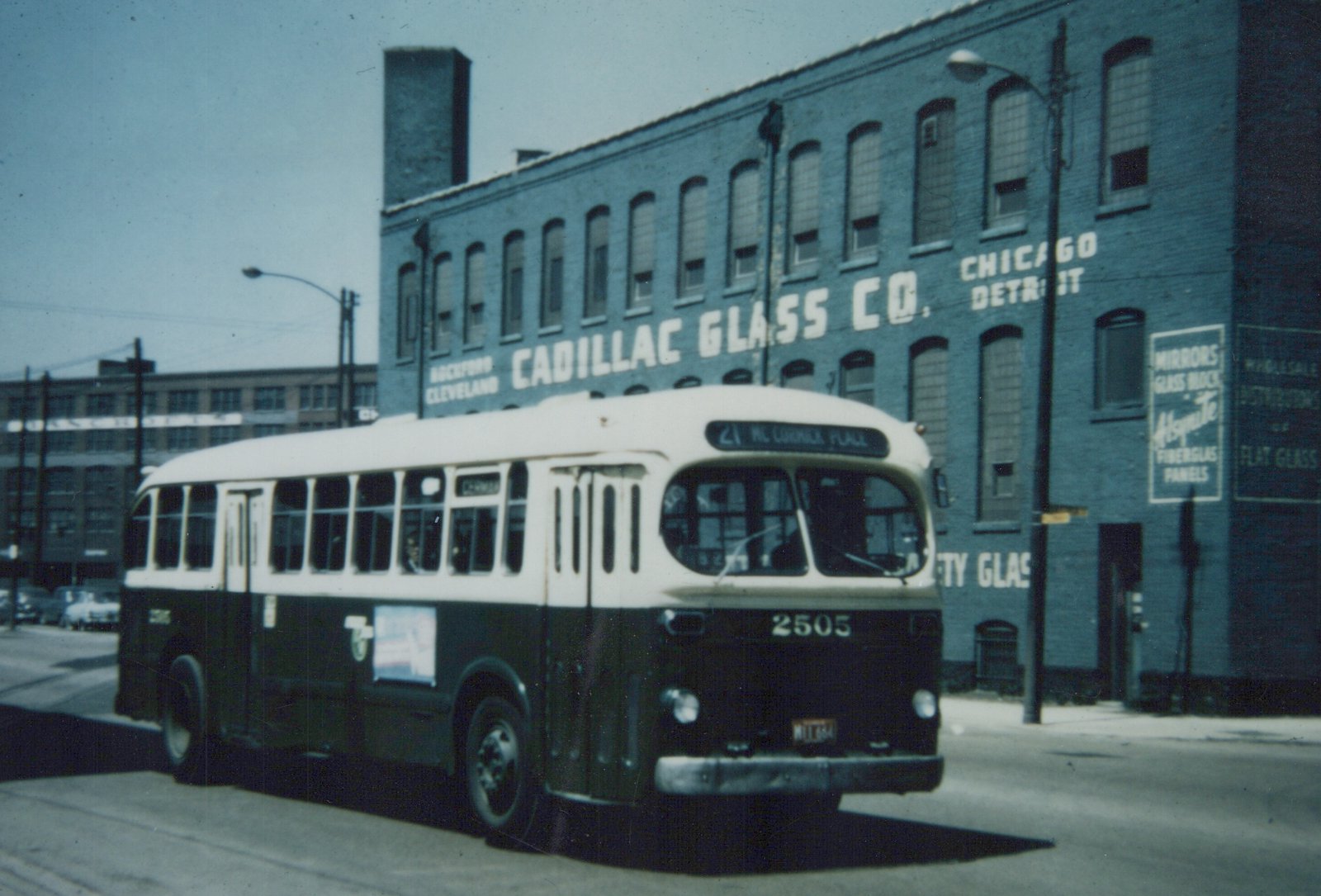 A vintage motor bus with a cream top and dark lower body, displaying route 21 to McCormick Place, is shown on a city street in front of a large brick building with 'Cadillac Glass Co.' painted on its side.