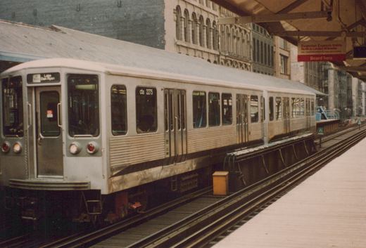 A silver Chicago 'L' car, numbered 2275, is stopped at an elevated station platform; a sign overhead reads 'Evanson Express trains North to Linden-Wilmette'.