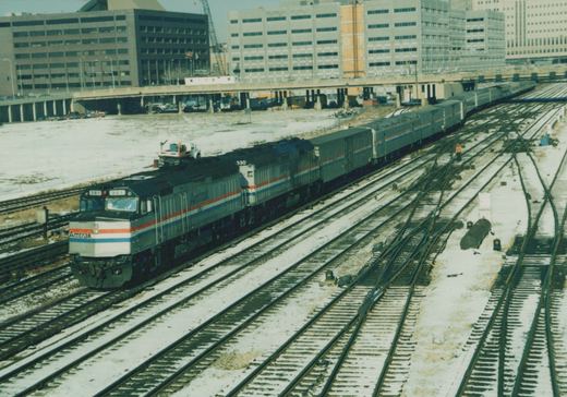 An Amtrak freight train with two locomotives and several passenger cars on a snowy day in a rail yard with city buildings in the background.