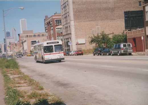 A white CTA bus with a red stripe, numbered 9972 and headed for Cottage Grove/94th, drives down a wide urban street lined with brick buildings and parked cars under a clear blue sky.