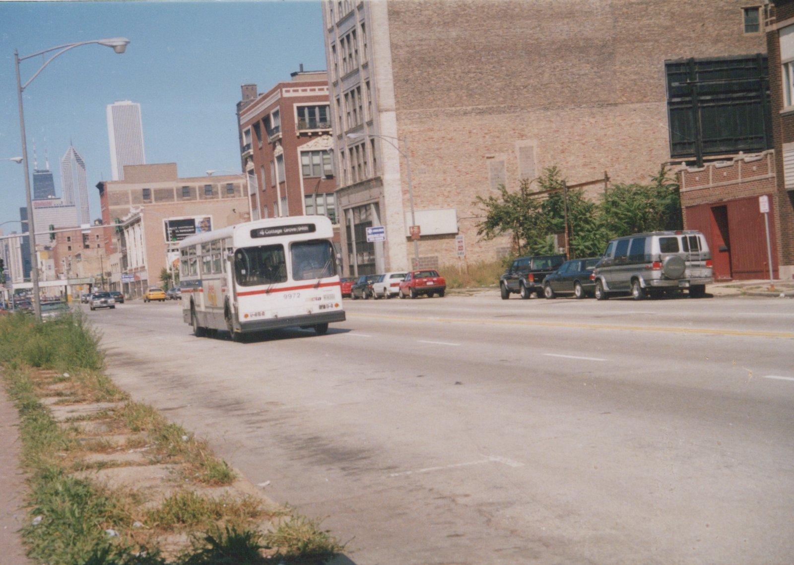 A white CTA bus with a red stripe, numbered 9972 and headed for Cottage Grove/94th, drives down a wide urban street lined with brick buildings and parked cars under a clear blue sky.