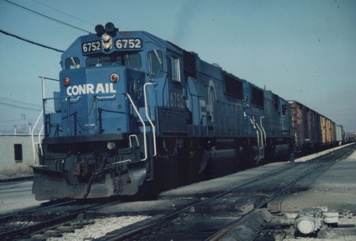 A blue Conrail locomotive numbered 6752 is pulling a long line of freight cars on railroad tracks next to a paved road, with a clear sky overhead.