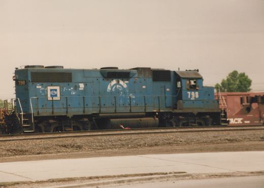A side view of a blue diesel locomotive with the number 798 and 'EDM Electric Motive' lettering, parked on tracks with a red freight car behind it.