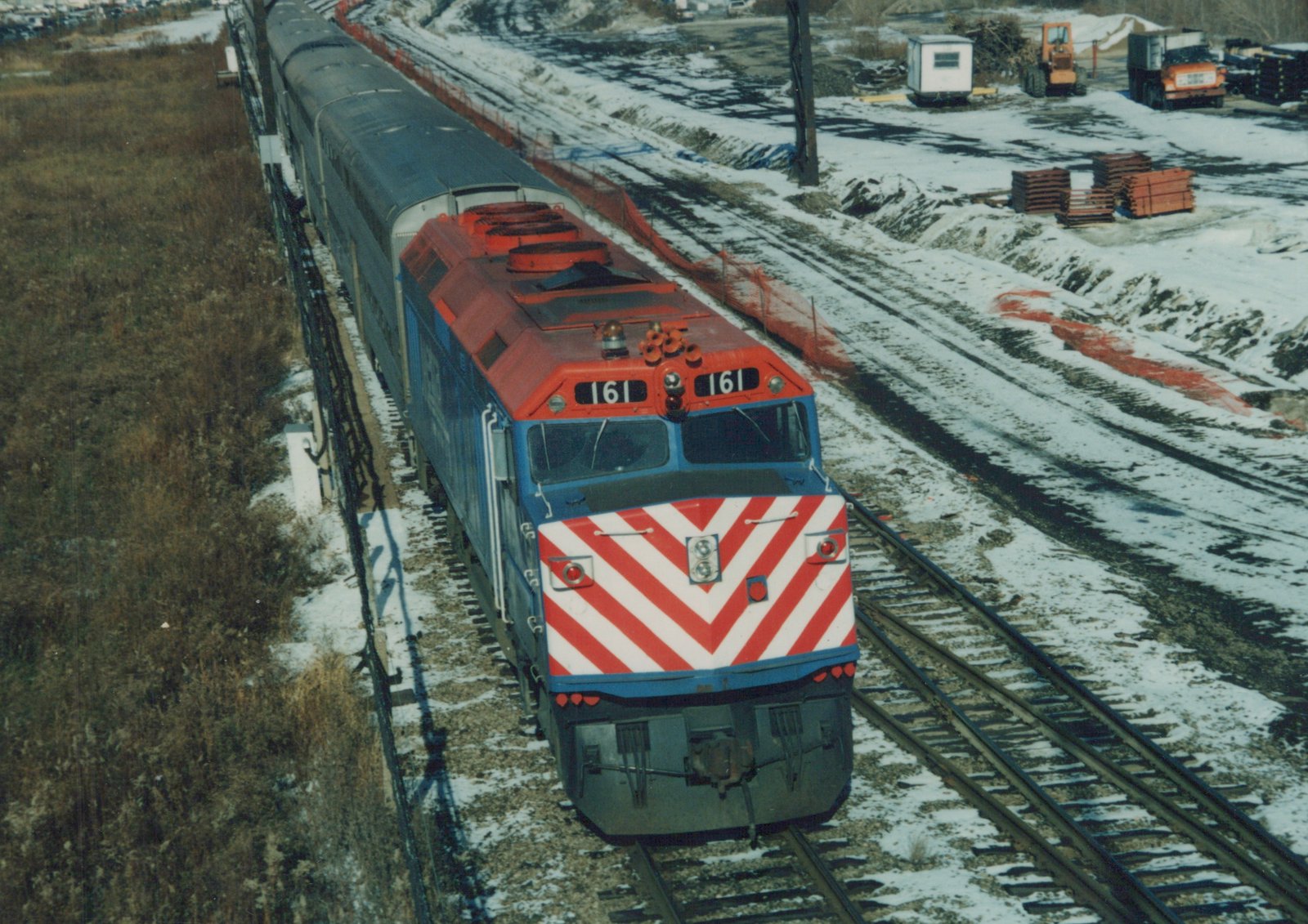 A blue and red diesel locomotive with a white and red chevron front, numbered 161, pulls a line of silver passenger cars along snow-covered tracks in winter.