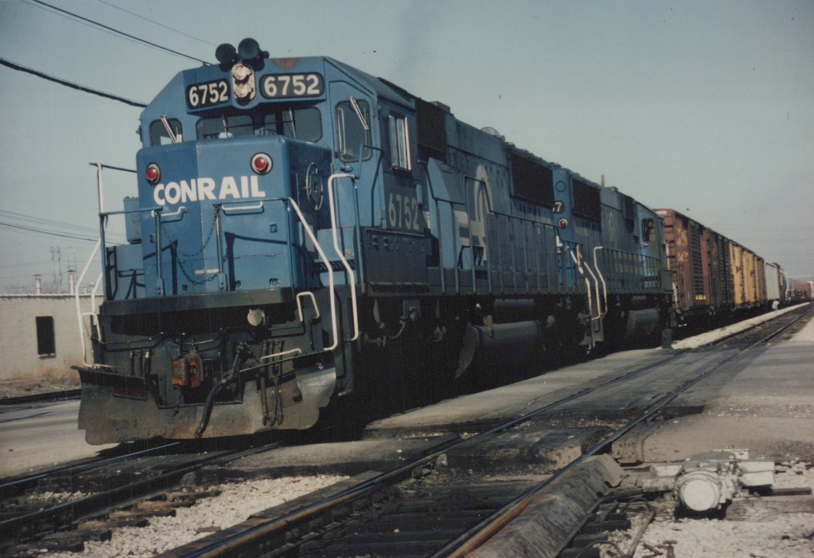A blue Conrail locomotive numbered 6752 is shown from a three-quarter front view, pulling a line of freight cars on a railroad track.