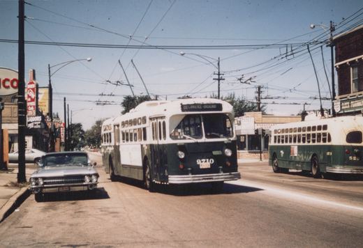 Two green and white Chicago Transit Authority trolleybuses on a city street under a network of overhead wires, with a 1960s car parked nearby and storefronts lining the street.
