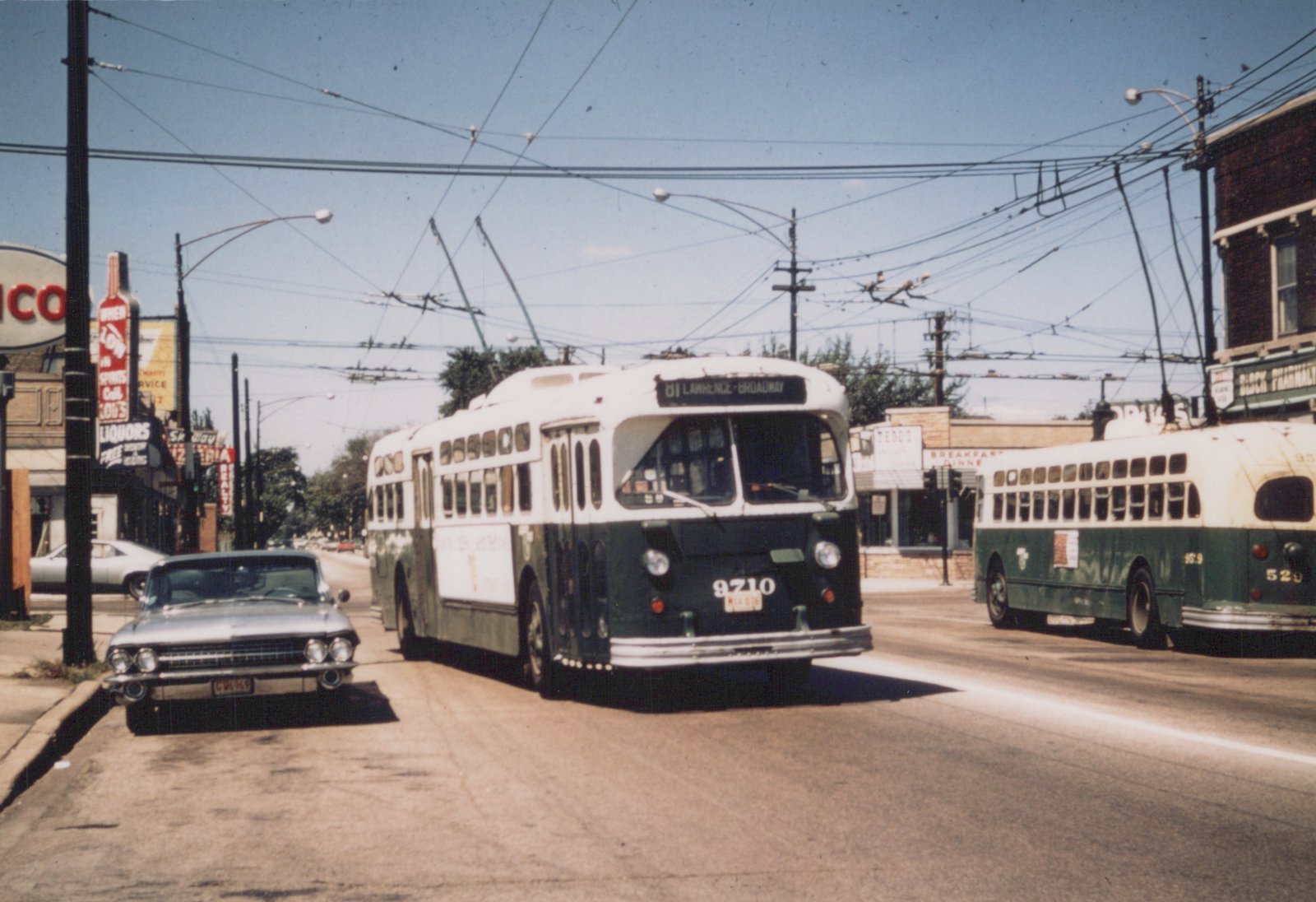 Two green and white Chicago Transit Authority trolleybuses on a city street under a network of overhead wires, with a 1960s car parked nearby and storefronts lining the street.