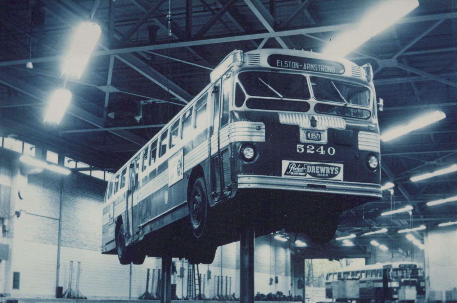 A vintage black and white photo of a city bus with the route ELSTON-ARMSTRONG on its sign, parked on a lift inside a large garage with high ceilings and fluorescent lights.