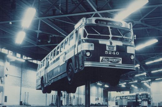 A vintage black and white photo of a city bus with the route ELSTON-ARMSTRONG on its sign, parked on a lift inside a large garage with high ceilings and fluorescent lights.