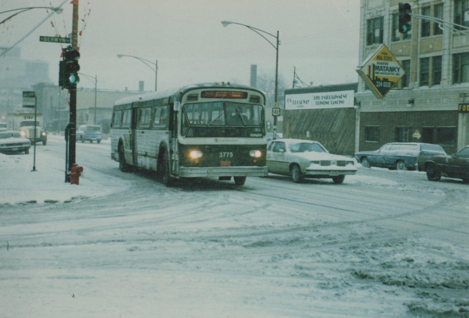 A white and green Chicago Transit Authority bus drives down a snow-covered street next to parked cars and storefronts in Berwyn, Illinois.