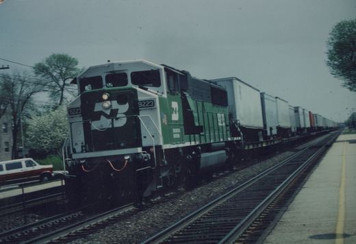 A green and white Burlington Northern diesel locomotive, number 9223, pulls a long line of boxcars past a grade crossing on a cloudy spring day.