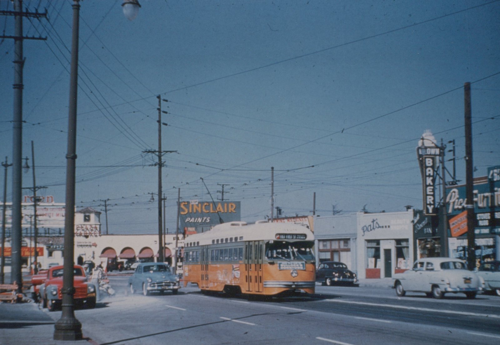 A yellow and cream Pacific Electric PCC streetcar with the number 28 on its front is driving down a busy street lined with 1950s cars and businesses like Sinclair Paints and Downtown Bakery.