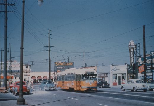 A yellow and cream Pacific Electric PCC streetcar with the number 28 on its front is driving down a busy street lined with 1950s cars and businesses like Sinclair Paints and Downtown Bakery.