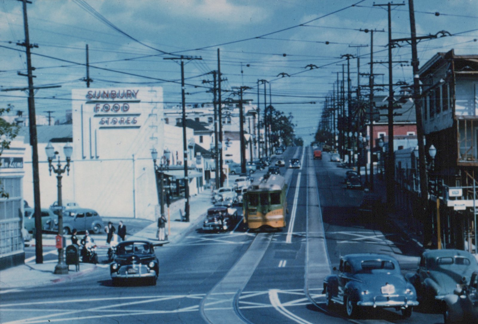 A vintage green and cream streetcar on a city street with 1940s cars, pedestrians, and a Sunbury Food Stores sign visible in the background.