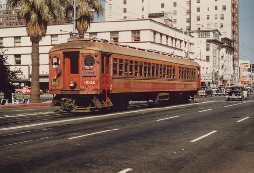 A vintage red interurban streetcar with the number 1541 and 'LONG BEACH LIMITED' written on its front travels down a city street lined with palm trees and 1950s cars.