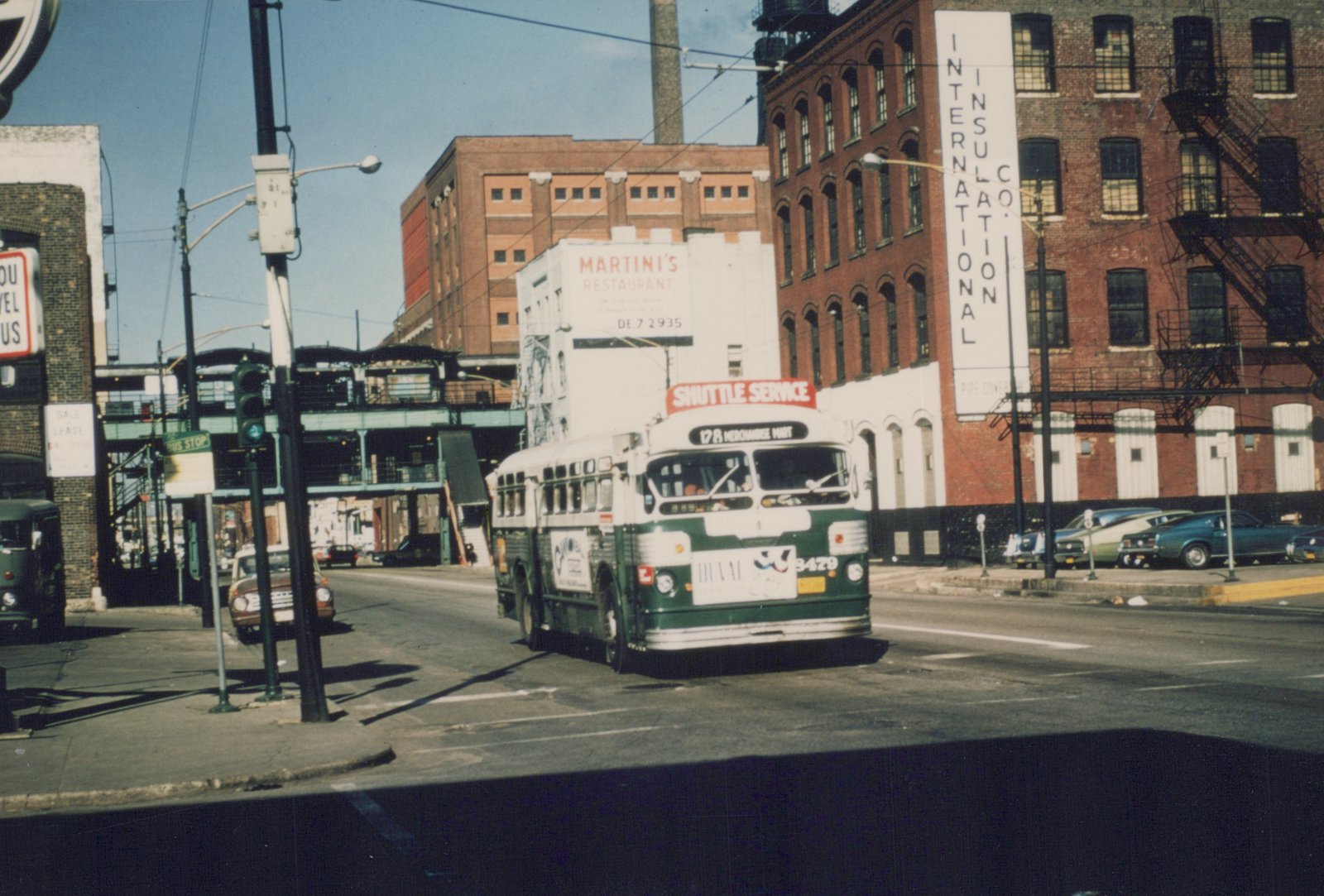 A white and green CTA bus labeled '128 MERCHANDISE MART' and 'SHUTTLE SERVICE' drives down a city street under the 'L' tracks, with brick buildings and parked cars nearby.