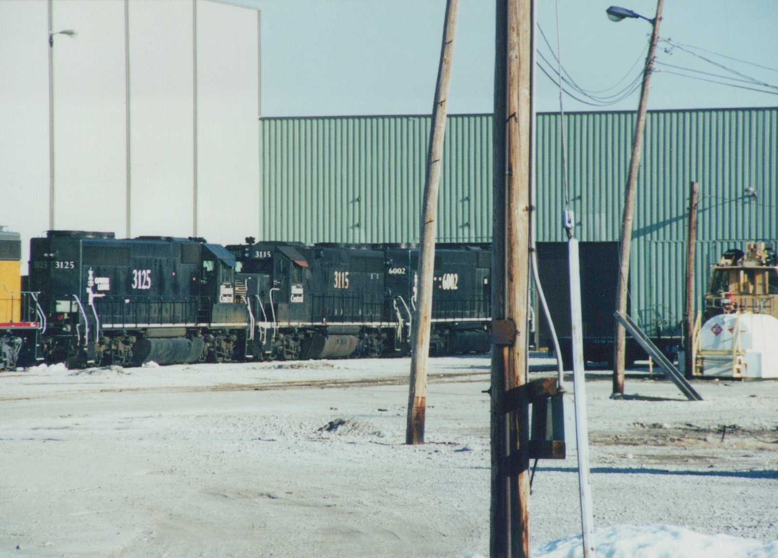 Three black diesel locomotives with Illinois Central lettering, numbered 3125, 3115, and 6002, are parked in a snowy rail yard, with industrial buildings and wooden utility poles in the background.