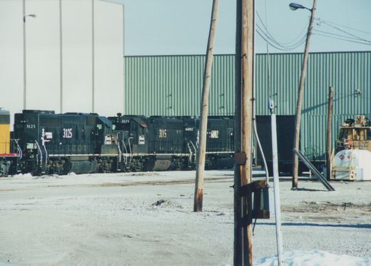 Three black diesel locomotives with Illinois Central lettering, numbered 3125, 3115, and 6002, are parked in a snowy rail yard, with industrial buildings and wooden utility poles in the background.
