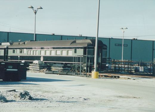 An Illinois Central passenger car in a snowy railroad yard with a green building in the background and a truck parked nearby.
