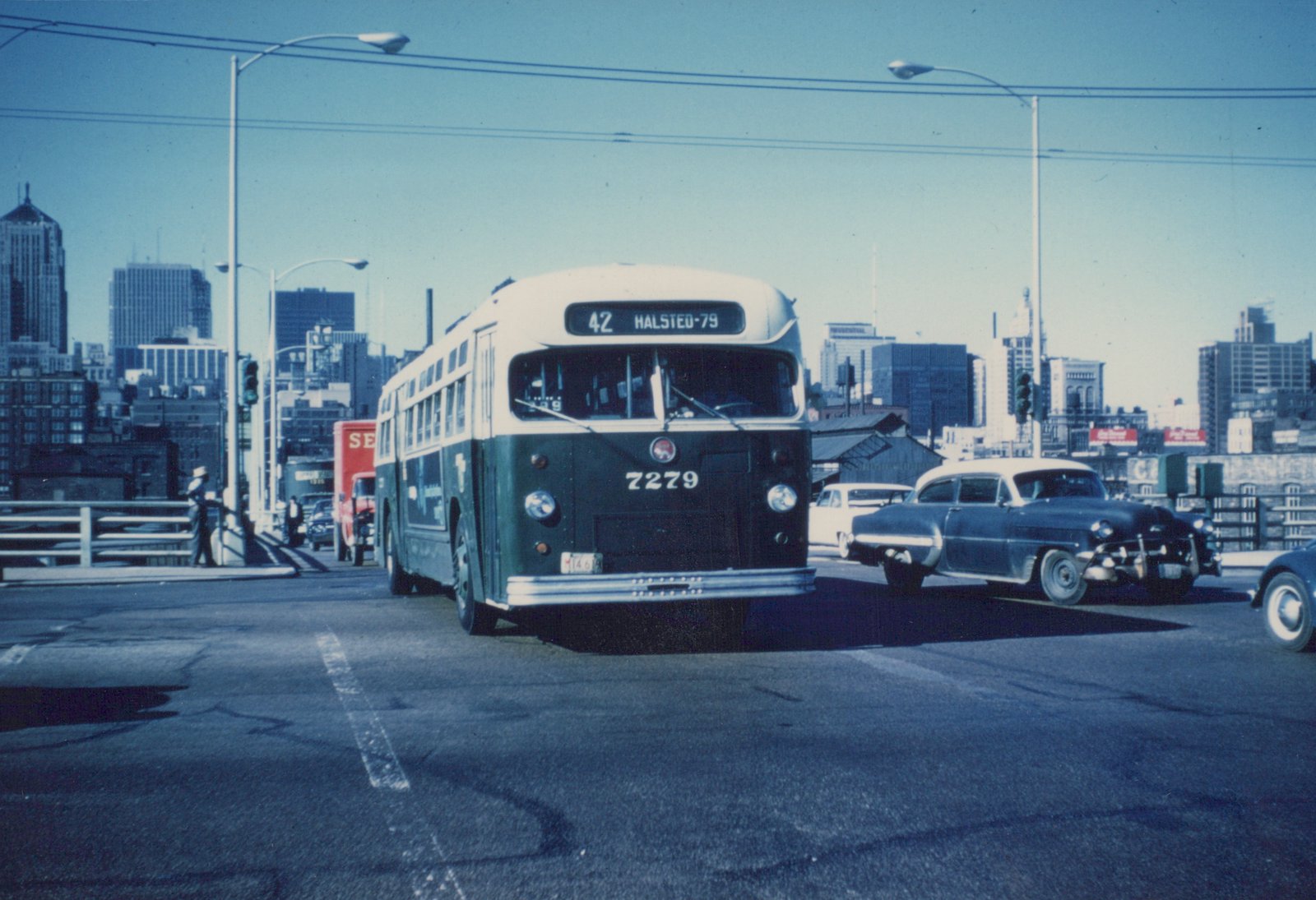 A dark green and cream CTA bus with the number 7279 on its front is stopped on a bridge with a city skyline in the background.