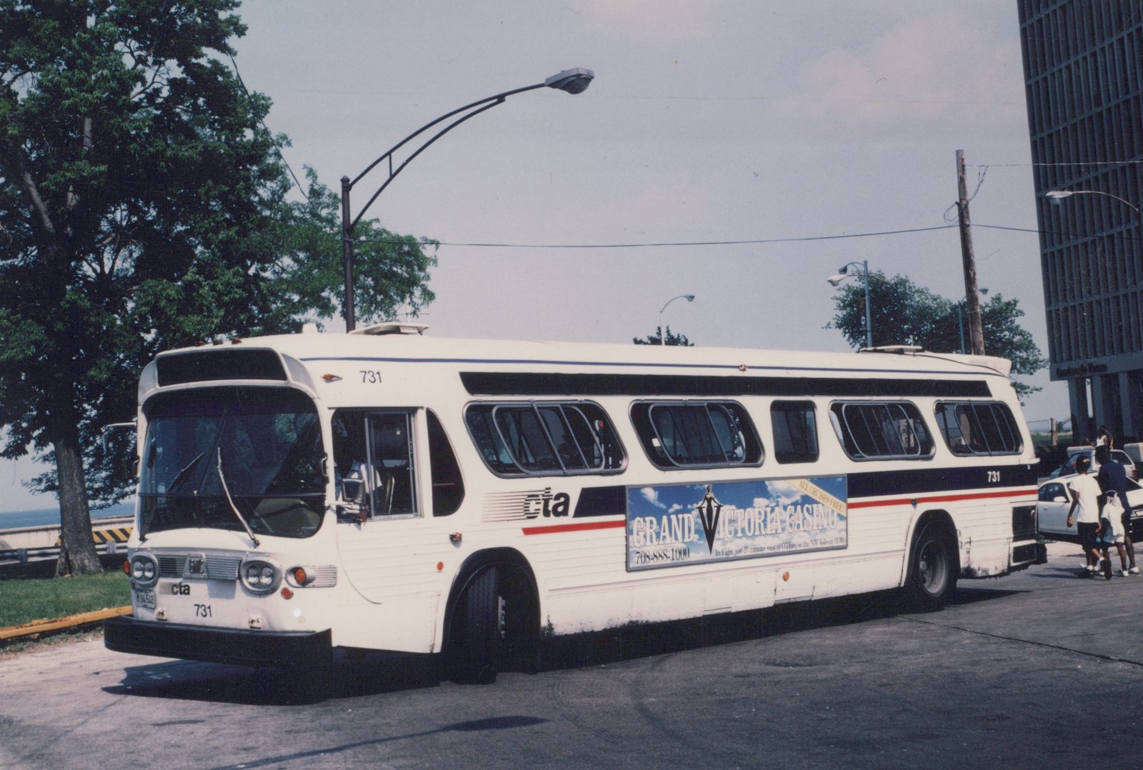 Side view of a white CTA bus with number 731 and a Grand Victoria Casino ad, parked on a street with pedestrians and a tall building in the background.