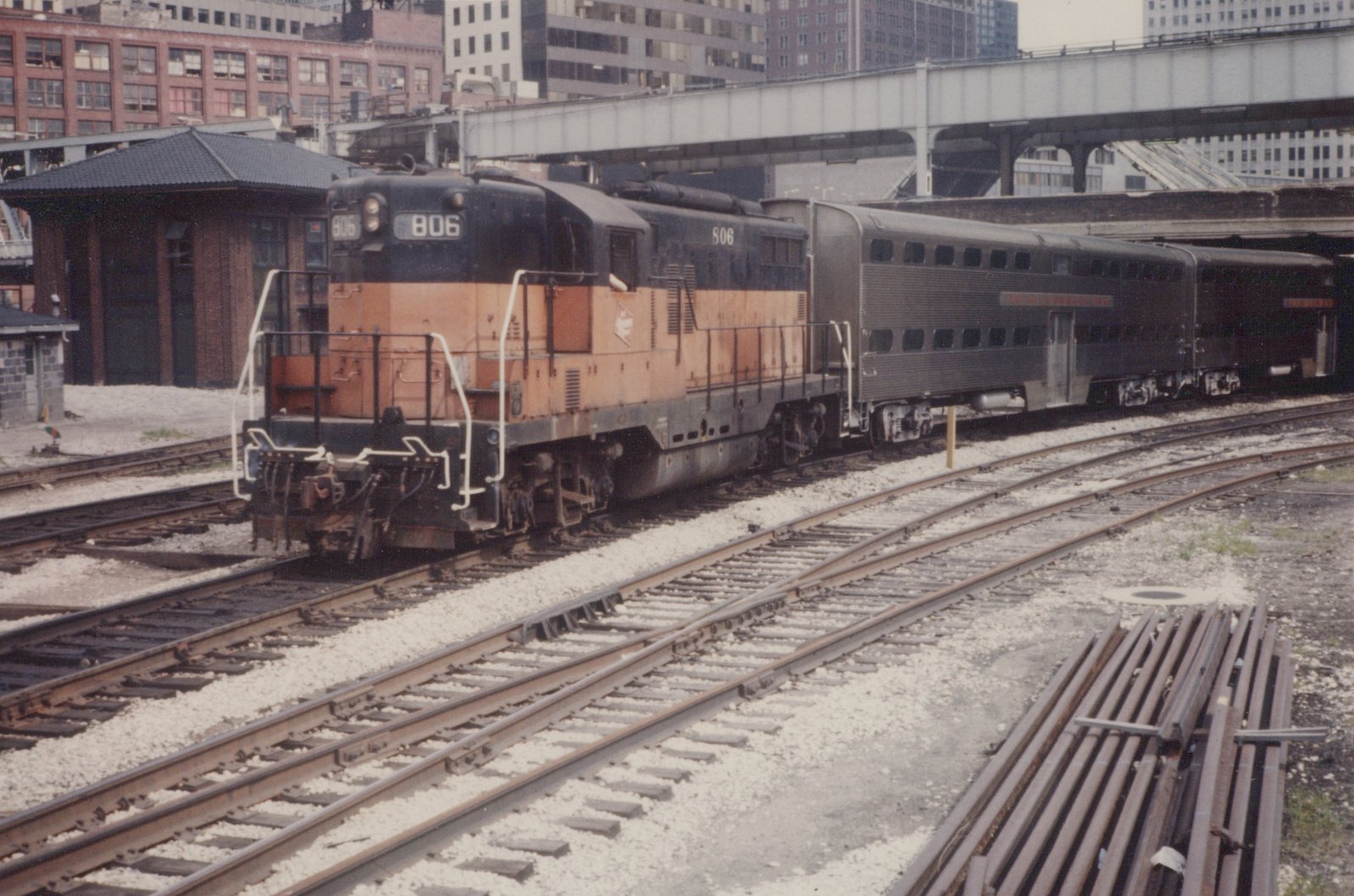 A black and orange diesel locomotive numbered 806 pulls two gray passenger cars on multiple tracks in a rail yard, with buildings and an elevated highway bridge in the background.